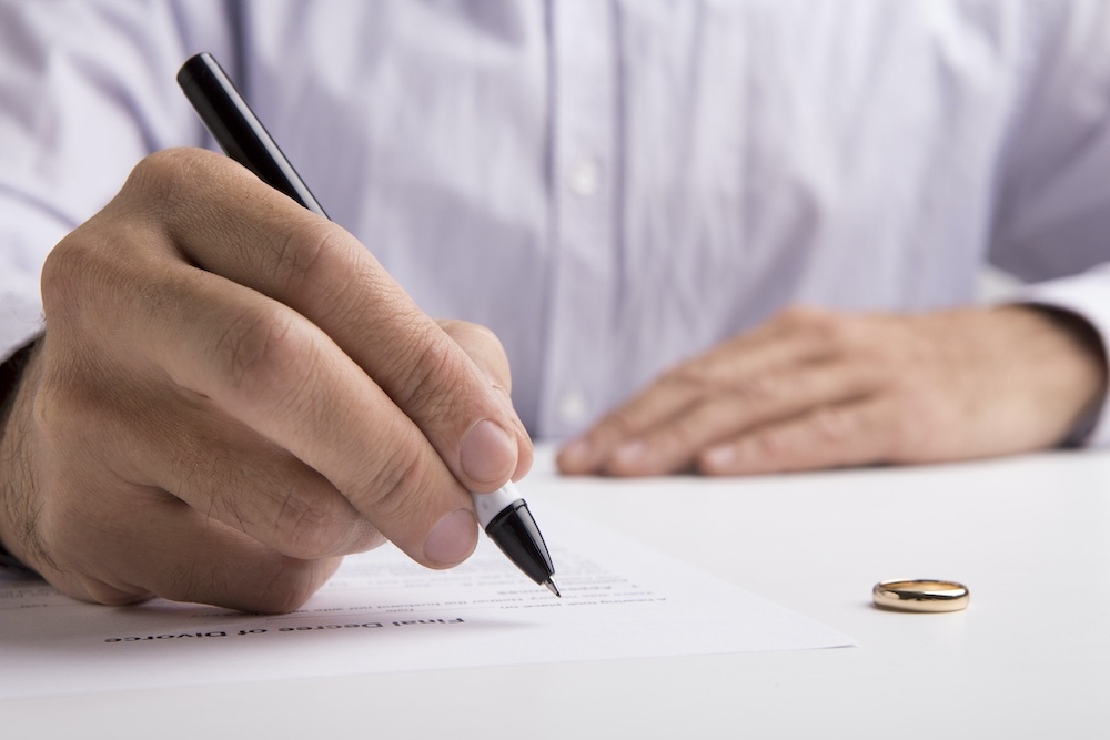 Man signing divorce papers with wedding ring on table